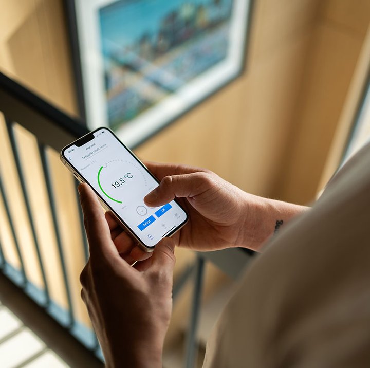 Person holder smarttelefon med app som viser temperaturinnstilling på 19,5 °C i et moderne hjem