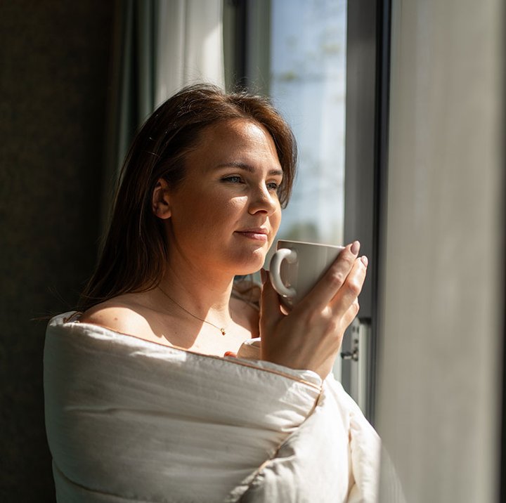 Person står ved vindu i bolig og drikker kaffe i dagslys, med rolig morgenstemning og naturlig lys.