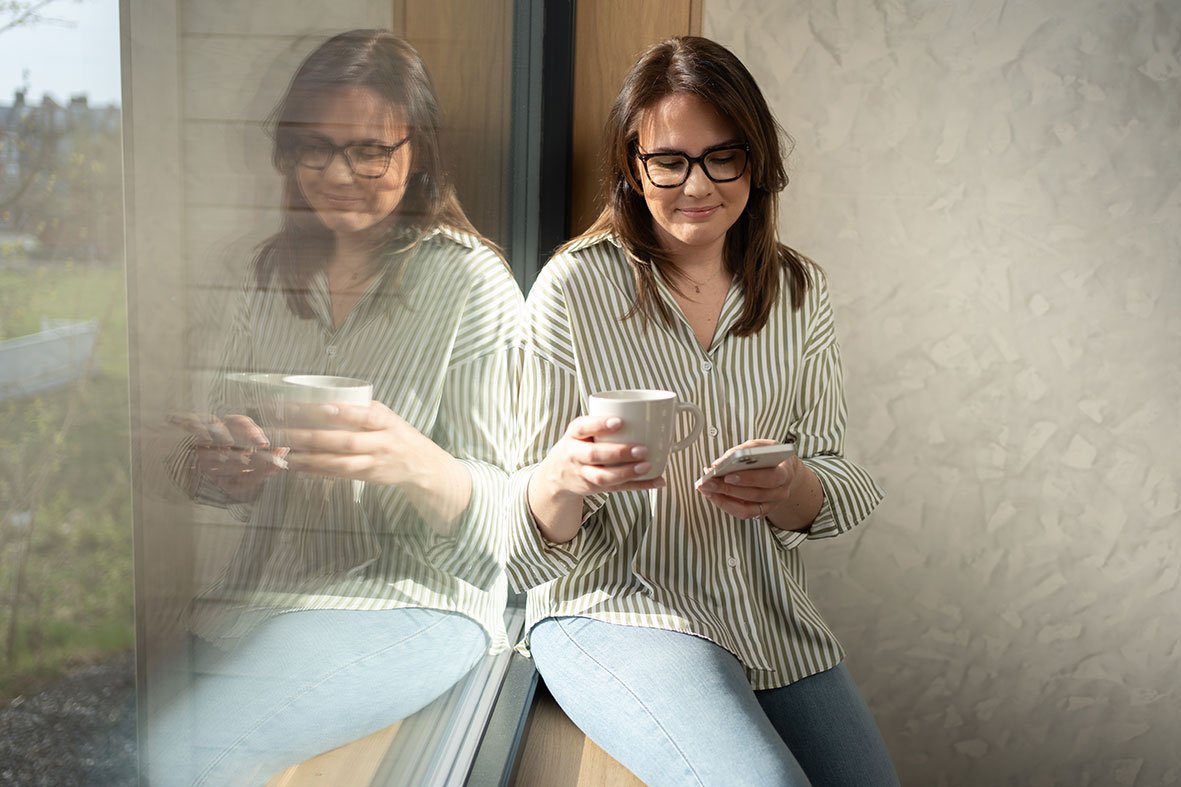 Person som bruker Flexit GO-appen på mobil for å styre ventilasjon og inneklima hjemme, mens de sitter ved et vindu.