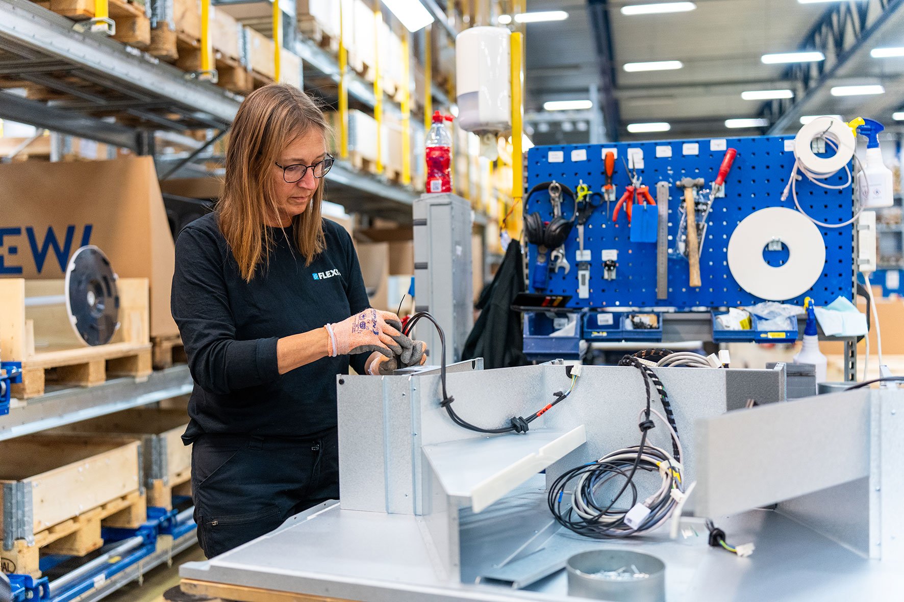 Ansatt hos Flexit smiler mens hun arbeider med montering av ventilasjonskomponent i produksjonsmiljø.