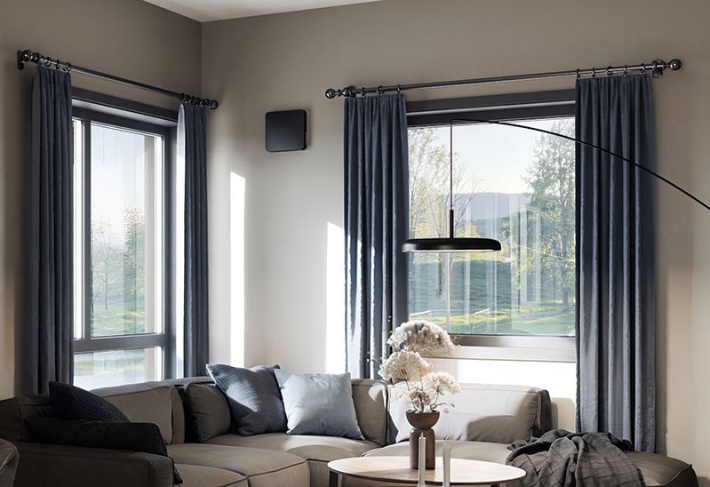 Romventilator montert høyt på vegg i stue med sofa, store vinduer og gardiner.